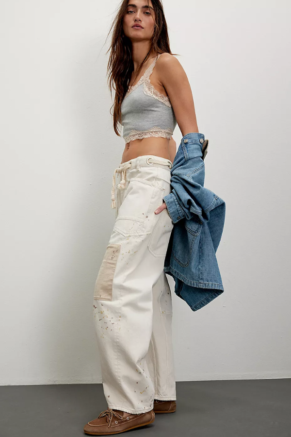 Woman wearing a gray crop top, white pants, and brown shoes, holding a denim jacket against a plain background.